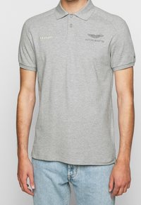 Šedé polo tričko vyrobené z bavlny, s límečkem, krátkými rukávy a vyšitými logy "HACKETT" a "ASTON MARTIN" v kontrastních barvách.