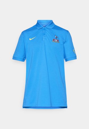 Polo azul de Nike con swoosh amarillo, escudo del Tottenham Hotspur en el pecho y logo en la manga derecha, exhibido sobre un fondo blanco.