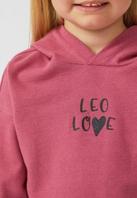 Felpa rosa con una texture morbida, caratterizzata da una stampa nera che riporta la scritta "LEO LOVE" accompagnata da un simbolo a forma di cuore. Cappuccio regolabile.