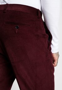 Burgundy manchesterbyxor med en bakficka som har en svart knapp och texturerad ribbad väv, i kombination med en vit långärmad skjorta.