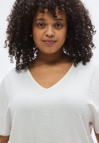 Λευκή μπλούζα με υφή, V-λαιμόκοψη και κοντά μανίκια. Διαθέτει διακριτικά κυματιστά σχέδια και απαλό, ελαφρύ ύφασμα.