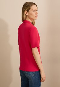 Fuksia lyhythihainen polo-paita, jossa on kauluspaita, sileä pinta, vartalonmyötäinen leikkaus, yhdistettyinä sinisiin farkkuihin, nähtynä takaa.