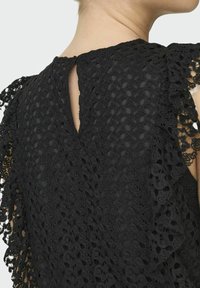 Top in pizzo nero con un design testurizzato e motivi ad intaglio. Presenta un'apertura a chiave sul retro e accenti in pizzo arricciato sulle spalle.