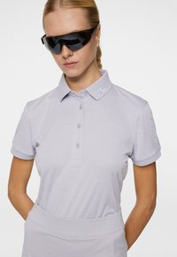 Ljusgrå polo med klassisk krage, tre knappar och accent på vänster ärm. Strukturen har en subtil lyster. Svarta solglasögon.