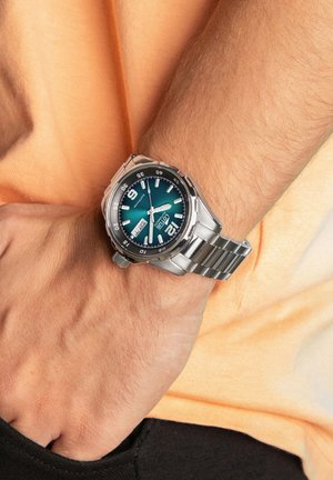Reloj de metal plateado con una esfera verde azulado, con marcadores de horas blancos y grandes números en las posiciones 9 y 12, unido a una correa de acero inoxidable.