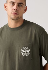 Μπλουζάκι από βαμβάκι σε ελιά πράσινο με κοντά μανίκια, που διαθέτει τσεπάκι στο στήθος και λευκό στρογγυλό λογότυπο. Ομαλή υφή.