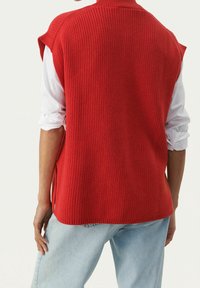 Rode gebreide vest met een geribbelde textuur, hoge halslijn en zijsplitsingen. Draag over een witte langemouwen shirt en gecombineerd met lichtblauwe jeans.