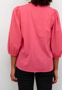 Ragazza vista di spalle che indossa un blouse rosa testurizzato con maniche a sbuffo a tre quarti e pantaloni neri su uno sfondo semplice.