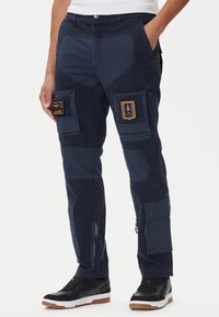 Pantaloni cargo navy con molte tasche, toppe di tessuto e cerniere; presenta distintivi con emblemi e texture a contrasto. Sneakers nere completano il look.