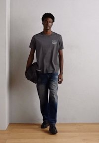 Grijze katoenen T-shirt met witte tekst, blauwe spijkerbroek en zwarte schoenen. Houdt een donkergrijze tas vast en staat tegen een lichte muur.