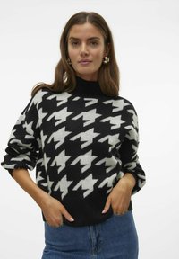 Juodai baltas houndstooth rašto megztinis su aukštu kaklu, laisvu siluetu ir mezginio tekstūra. Suderintas su mėlynomis džinsinėmis kelnėmis.