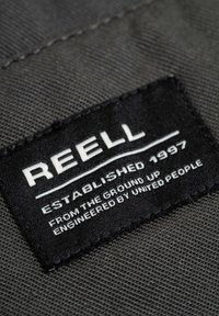 Black fabric label with white text reading "REELL ESTABLISHED 1997 FROM THE GROUND UP ENGINEERED BY UNITED PEOPLE." Text is contrasted and bold.