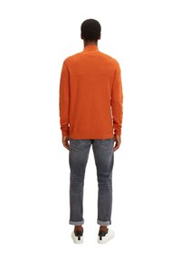 Pull en tricot orange avec un col montant, des manches longues et une texture côtelée, associé à un jean gris et des baskets blanches avec des accents noirs.