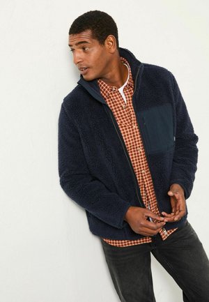 Marineblaue Fleece-Zip-Jacke mit Kapuze und strukturiertem Material, ausgestattet mit einer Brusttasche. Tragend über einem karierten Hemd und schwarzen Jeans.