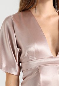 Sukně z saténu v světle růžové barvě s hlubokým výstřihem do V, krátkými rukávy a přiléhavým pasem. Má hladkou texturu a jemný lesk.