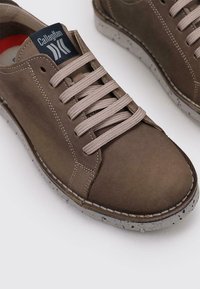 Zapatilla de piel marrón con parte superior texturizada, cordones planos y suela de goma moteada. Presenta un parche con el logotipo en la lengüeta.