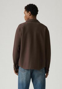 Brązowa teksturowana koszula z kołnierzykiem, długimi rękawami i zapinanymi na guziki mankietami, noszona na niebieskich, spranych dżinsach. Czysty i minimalistyczny design.