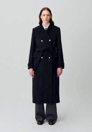 Donna che indossa un lungo cappotto nero con cintura e bottoni dorati, camicia bianca, cravatta a righe, pantaloni grigi a righe e scarpe marrone scuro, in piedi.