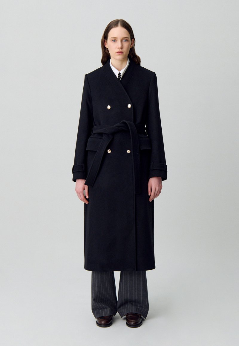 Donna che indossa un lungo cappotto nero con cintura e bottoni dorati, camicia bianca, cravatta a righe, pantaloni grigi a righe e scarpe marrone scuro, in piedi.
