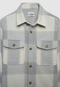 Chemise à carreaux gris clair et crème avec col, deux poches poitrine et boutons dorés. Fabriquée en tissu doux, elle présente une coupe confortable.