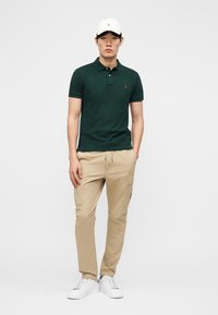 Uomo in piedi che indossa una polo verde scuro, pantaloni cargo beige, sneakers bianche e cappellino da baseball bianco su sfondo grigio chiaro uniforme.