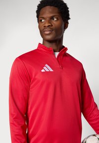 Κόκκινο αθλητικό μακρυμάνικο πουκάμισο με φερμουάρ και ψηλό γιακά, με λευκό λογότυπο Adidas στο αριστερό μέρος του στήθους. Λεία υφή.