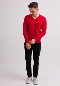 Maglione rosso a V in tessuto morbido, abbinato a pantaloni neri aderenti e sneaker bianche, in piedi su uno sfondo chiaro.