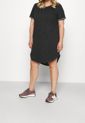 Femme portant une robe noire à manches courtes avec un imprimé léopard subtil et des baskets mauves New Balance, debout sur un sol gris.