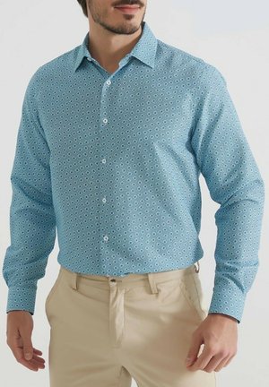 Chemise bleu clair à boutons avec un motif circulaire en bleu foncé. Manches longues avec fermetures à boutons, associée à un pantalon beige.