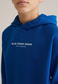 Kék kapucnis pulóver puha anyagból, húzózsinóros kapucnival. Elöl fehér szöveg: "BLUE RIDGE DENIM STAY FEARLESS" található.