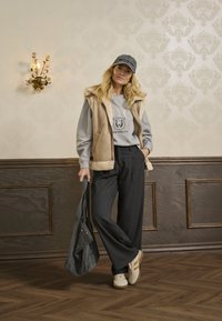 Tan vest met bontvoering over een grijs sweatshirt met 'NYC'-print, zwarte broek met wijde pijpen, grijze tas en witte sneakers met tan-strepen.