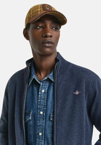 Marinblå stickad tröja med dragkedja över en ljusblå denimskjorta. Kepsen är mönstrad i gult och brunt med en brun läderlogga.