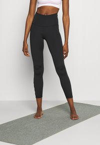 Leggings neri a vita alta realizzati in tessuto elasticizzato, design aderente e texture liscia, abbinati a un top corto rosa chiaro.