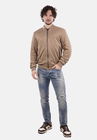 Giacca bomber in suede beige con zip frontale, tasche laterali, abbinata a jeans chiari strappati e sneakers marroni con dettagli bianchi.