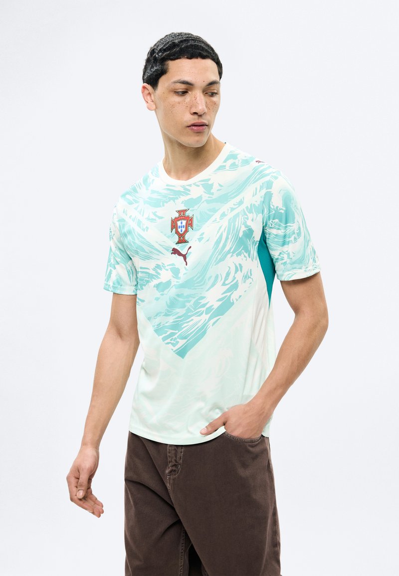 Mladý muž s krátkymi kučeravými vlasmi, oblečený v svetlom akvamarínovom športovom drese s portugalským futbalovým erbom a hnedými nohavicami.