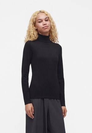 Jeune personne aux longs cheveux blonds bouclés portant un pull à col roulé noir et un pantalon ample gris foncé sur un fond clair uni.