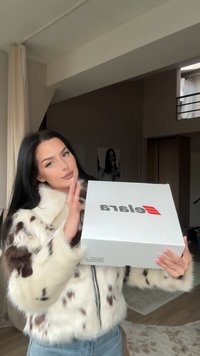 Vit låda med svart text som säger "elara," hållen framför en person. Personen har på sig en fuskpälsjacka med ett prickigt mönster.