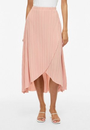 Kvinne som har på seg et høyt liv, plissert omslags-midiskjørt i myk rosa med hvite sandaler med stropper og hæl.