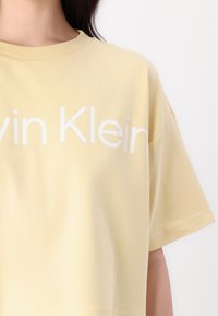 Gul bomullst-skjorte med rund hals, korte ermer og en stor hvit "Calvin Klein" logo over brystet. Glatt tekstur.