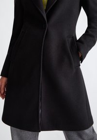 Cappotto di lana nero con design sartoriale, dotato di chiusura a bottone singolo, tasche laterali e orlo leggermente svasato. Tessuto con texture.