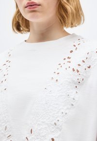 Κοντινό πλάνο ατόμου που φοράει λευκό τοπ με περίτεχνη δαντέλα με λουλουδένια σχέδια στους ώμους και το στήθος.