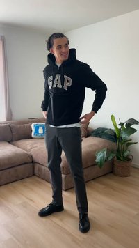 Mann trägt schwarzen GAP-Hoodie, graue Hose und schwarze Schuhe, steht im Wohnzimmer mit beigem Sofa und grünem Pflanze.