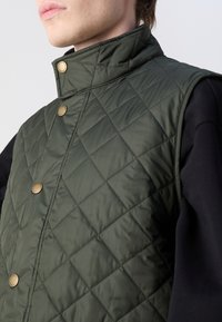 Gilet verde trapuntato con collo alto, caratterizzato da bottoni a pressione dorati. Il materiale sembra leggero con una texture liscia e un motivo a diamante.