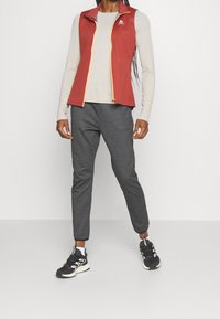 Röd ärmlös väst med gul dragkedja, ljusgrå långärmad tröja, grå smalbyxor och svarta sportskor med vita detaljer.