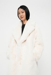 Cremefarvet kunstpelsjakke med bred, spidse krave, oversized silhuet og store sidelommer. Blød tekstur med subtile variationer.