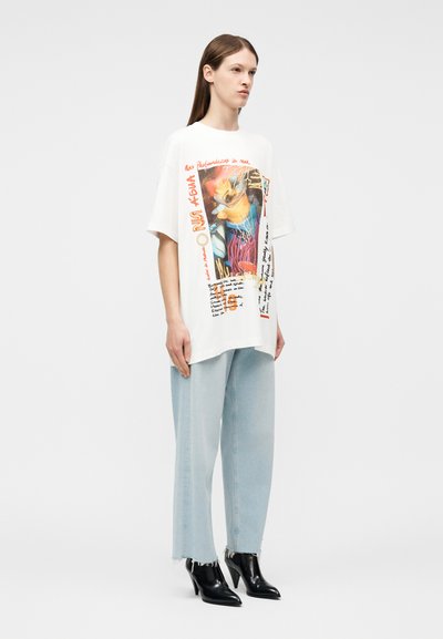 Νεαρή γυναίκα όρθια, φοράει λευκό φαρδύ T-shirt με πολύχρωμο σχέδιο, ανοιχτό μπλε φαρδιά τζιν και μαύρες μπότες με τακούνι μέχρι τον αστράγαλο.