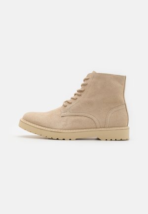 Beige ruskindsanker med rund tå, seks metaløjer og en tekstureret gummisål med dybe mønstre. Simpelt design.