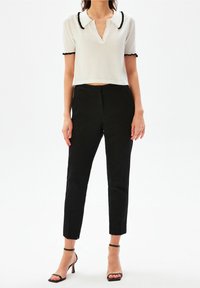 Valkoinen lyhythihainen cropped-toppi, jossa on musta aaltoileva kaulus, yhdistettynä mustiin slim-fit-housuihin ja mustiin korkokenkiin.