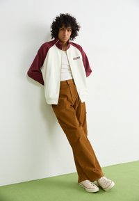 White and maroon zip-up jacket, white tank top, brown corduroy pants, and beige sneakers. Model leans against a wall on green floor.