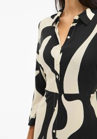 Črna in krem dolgrokavnik obleka z drznim abstraktnim vzorcem, ovratnikom in gladkim materialom.